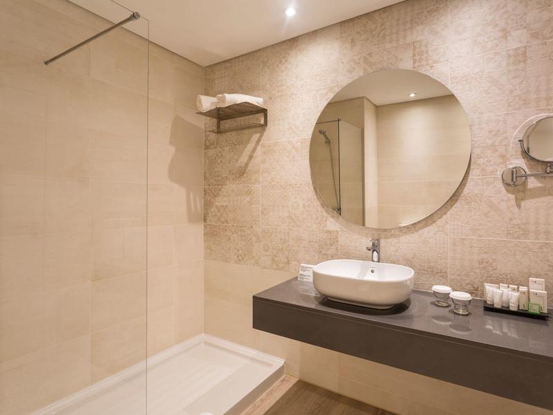 Salle de bain moderne avec douche à l’italienne, miroir rond et lavabo sur un comptoir sombre.