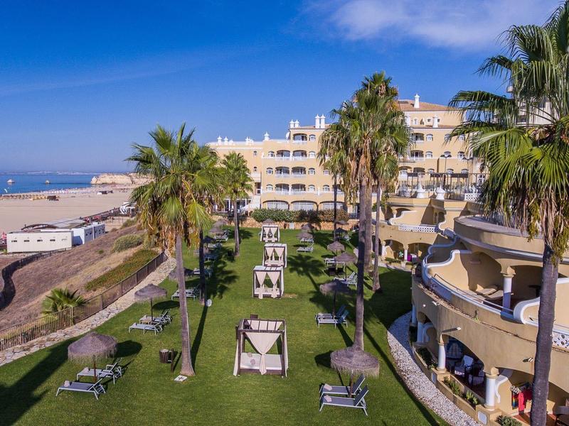 Vue sur un jardin d'hôtel avec des palmiers, des chaises longues et une vue sur la plage de sable et la mer.