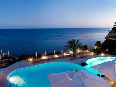 Espace piscine d'un hôtel au bord de la mer au coucher du soleil avec des palmiers éclairés et des chaises longues