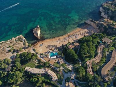 Vue aérienne d'une plage avec une eau claire, entourée de forêts verdoyantes et de bâtiments.