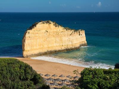 Formation rocheuse près d'une plage de sable avec parasols et mer bleue en arrière-plan