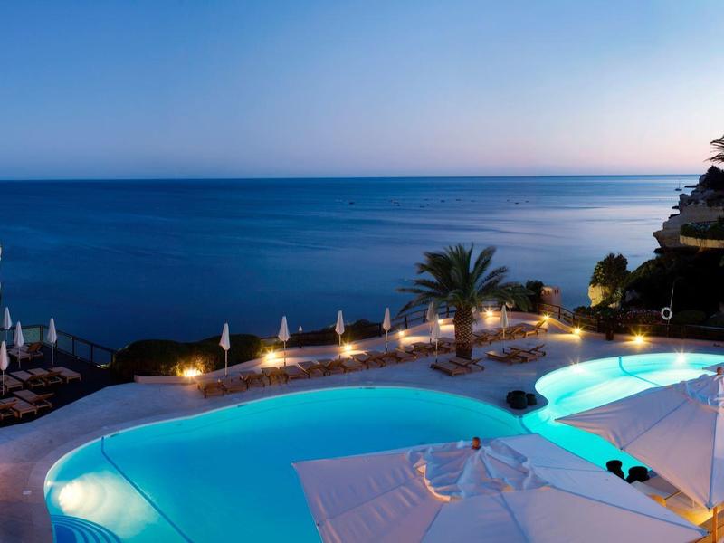 Espace piscine d'un hôtel au bord de la mer au coucher du soleil avec des palmiers éclairés et des chaises longues