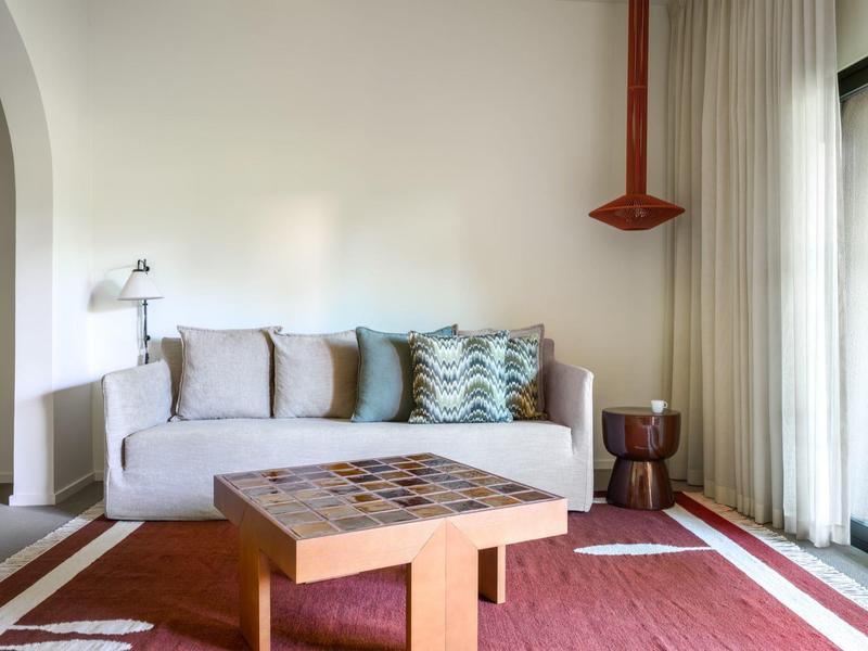 Salon moderne avec canapé gris, coussin coloré, table en bois et tapis rouge près de la fenêtre.