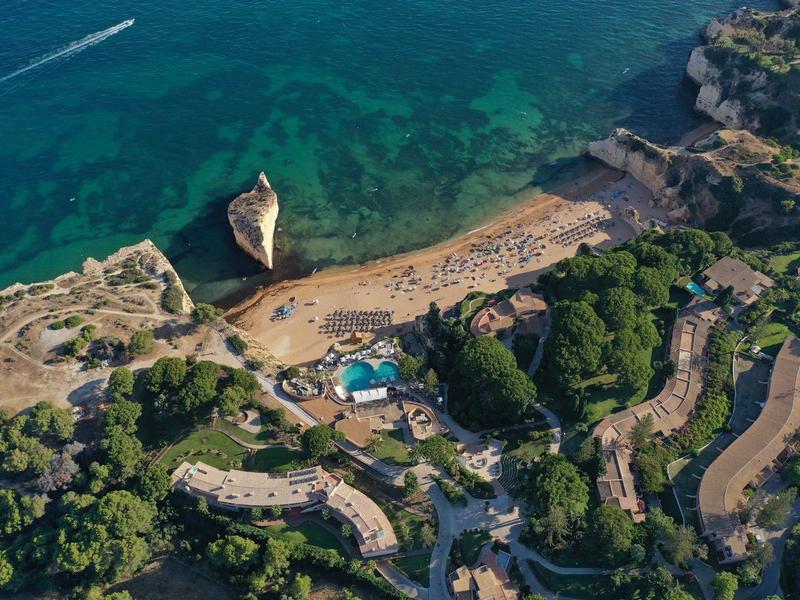 Vue aérienne d'une plage avec une eau claire, entourée de forêts verdoyantes et de bâtiments.