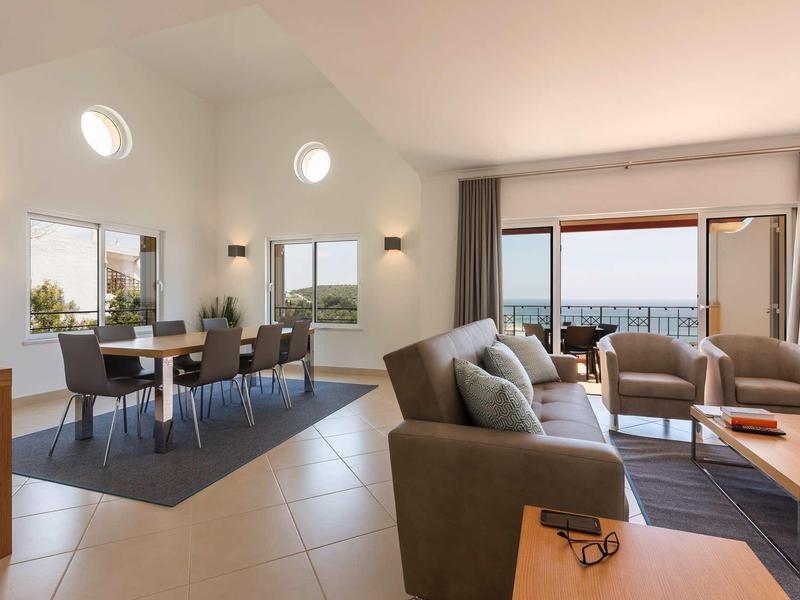 Salon lumineux avec canapé gris, coin repas et vue sur la mer à travers de grandes fenêtres et portes-fenêtres.