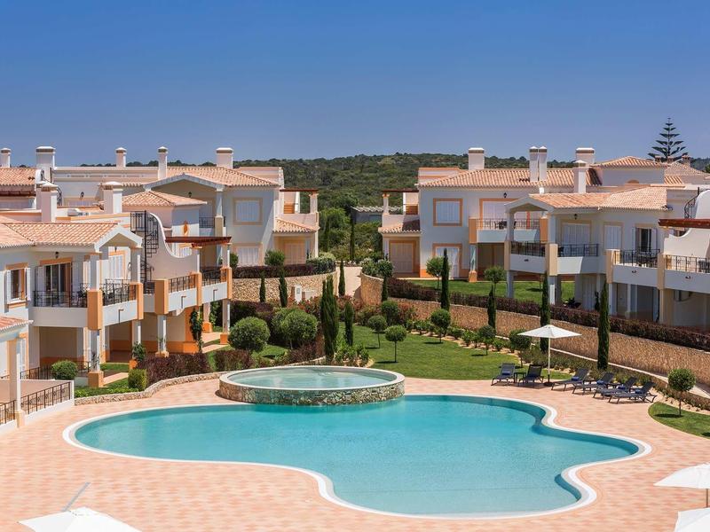 Grande piscine extérieure entourée de maisons de vacances modernes sous un ciel bleu clair.