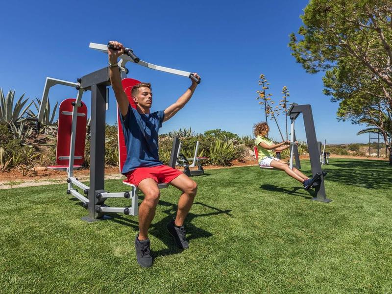 Deux personnes font de l'exercice en plein air sur des appareils de fitness sur une pelouse verte sous un ciel ensoleillé.
