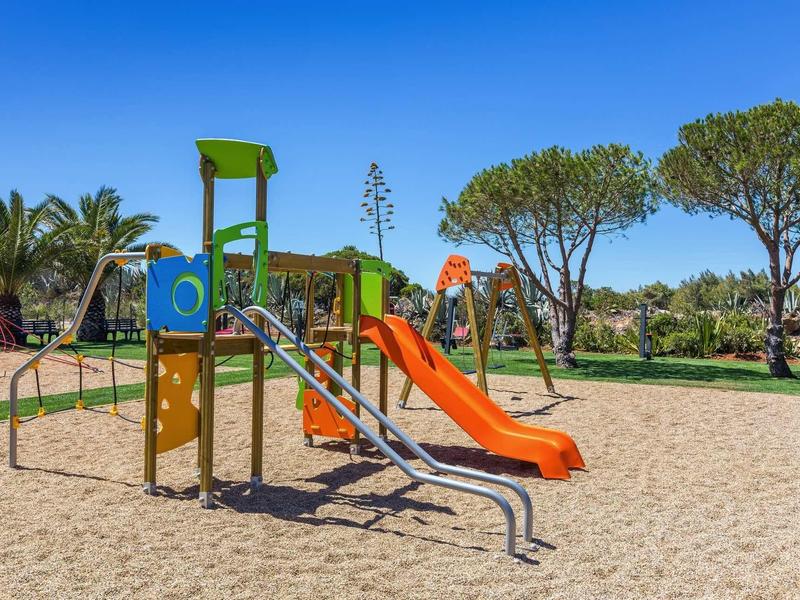 Aire de jeux pour enfants avec toboggan et structure d'escalade sur sol sablonneux sous un ciel dégagé.