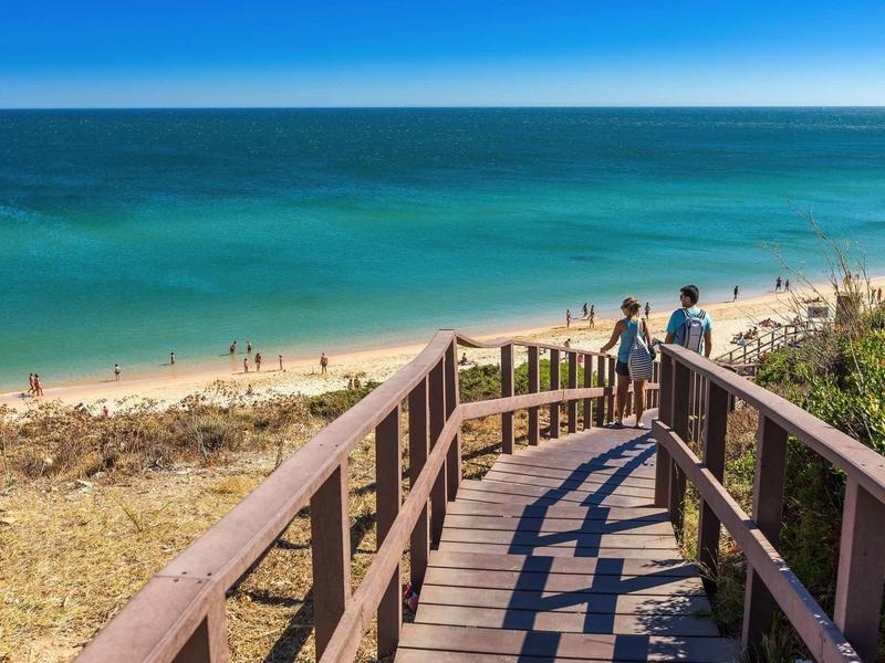 Passerelle en bois menant à une plage de sable avec mer bleue et ciel clair.
