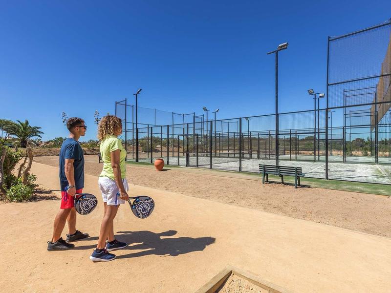 Deux personnes avec des raquettes marchent vers un court de padel clôturé sous un ciel bleu.