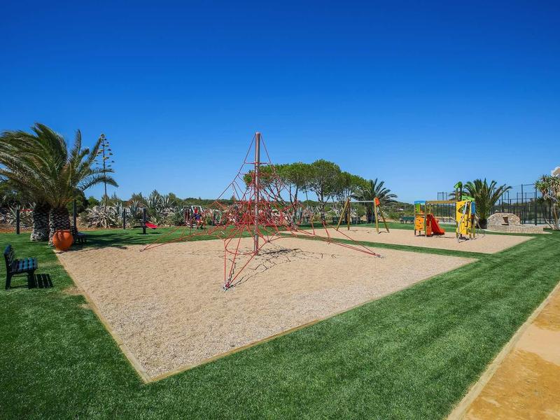 Aire de jeux avec zone de sable, structure d'escalade et équipements colorés dans un parc sous un ciel bleu clair.