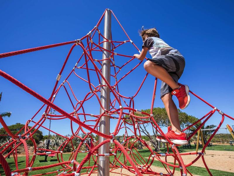 Un enfant grimpe sur une structure d'escalade en corde rouge dans une aire de jeux sous un ciel bleu clair.