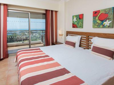 Tweepersoonskamer met witte bedden en rood gestreepte dekens voor balkon met uitzicht op zee.