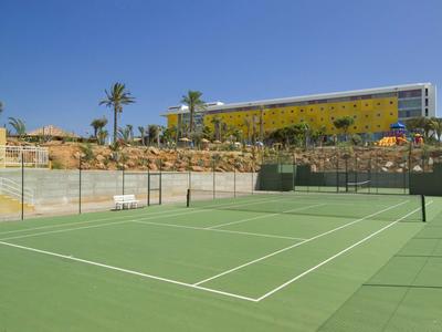 Tenniscourt mit grünem Belag vor Hotelgebäude und Palmen unter blauem Himmel.