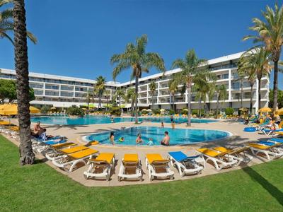 Großer runder Pool mit Sonnenliegen und Palmen vor einem Hotelgebäude im Sonnenschein.