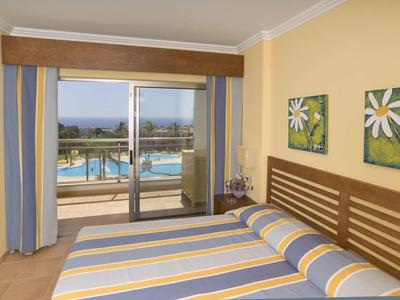 Slaapkamer met tweepersoonsbed en balkon met uitzicht op zwembad en zee in warme kleuren.