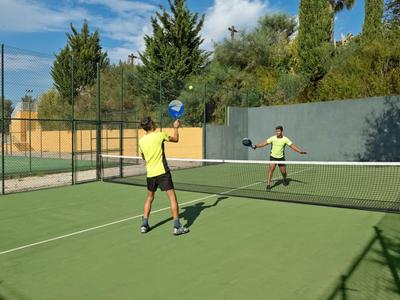 Twee mensen spelen padeltennis op een zonnige buitenbaan met groene omgeving.