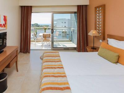 Hotelkamer met tweepersoonsbed, tv, bureau en balkon met zeezicht.