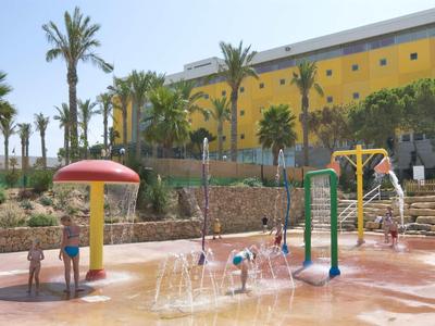 Water-speeltuin met fonteinen, glijbanen en palmbomen voor een geel gebouw.