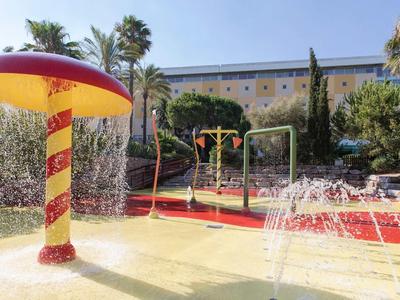 Kleurrijke waterspeeltuin met paddenstoelfontein en spuitend water onder blauwe lucht.