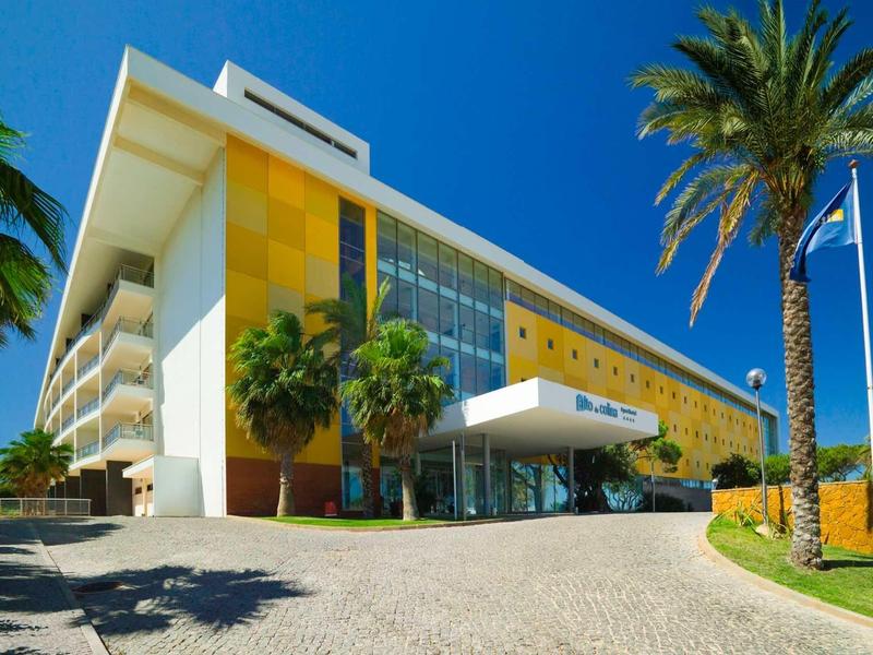 Modern hotel met gele gevel, palmbomen en een heldere blauwe lucht op de achtergrond.