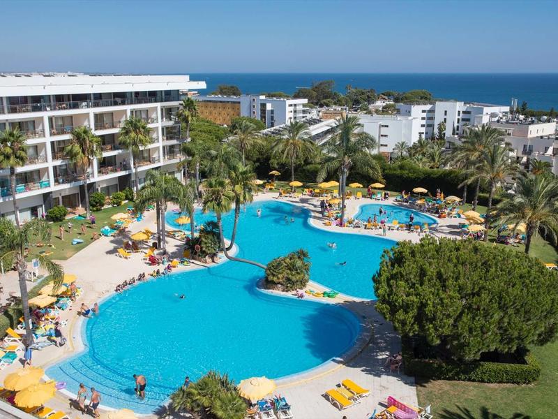 Groot hotelzwembad met parasols en ligstoelen, omgeven door een meerlaagse hotelgebouw en palmbomen, zee op de achtergrond