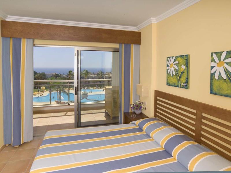 Slaapkamer met tweepersoonsbed en balkon met uitzicht op zwembad en zee in warme kleuren.