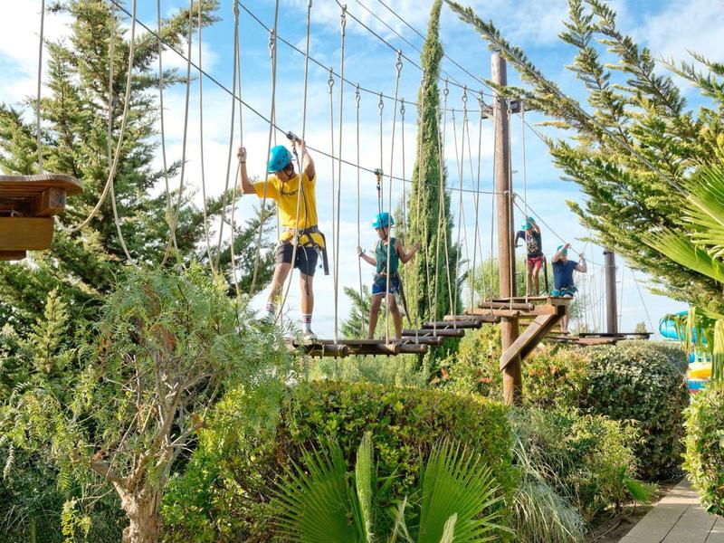 Mensen steken een touwenparcours over met veiligheidsbanden in een groene hotelomgeving bij heldere lucht.