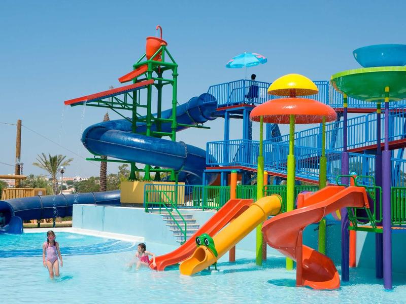 Kinderen spelen in het ondiepe zwembad met kleurrijke waterglijbanen en waterspeeltoestellen onder een zonnige hemel.