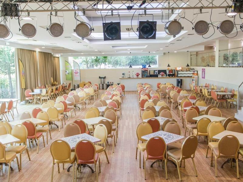 Grote evenementenhal met rijen tafels en stoelen, podium aan de voorkant met verlichting.