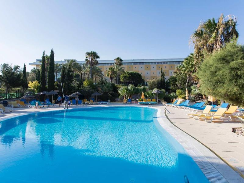 Groot buitenzwembad met ligstoelen en bomen in een hoteltuin op een zonnige dag.