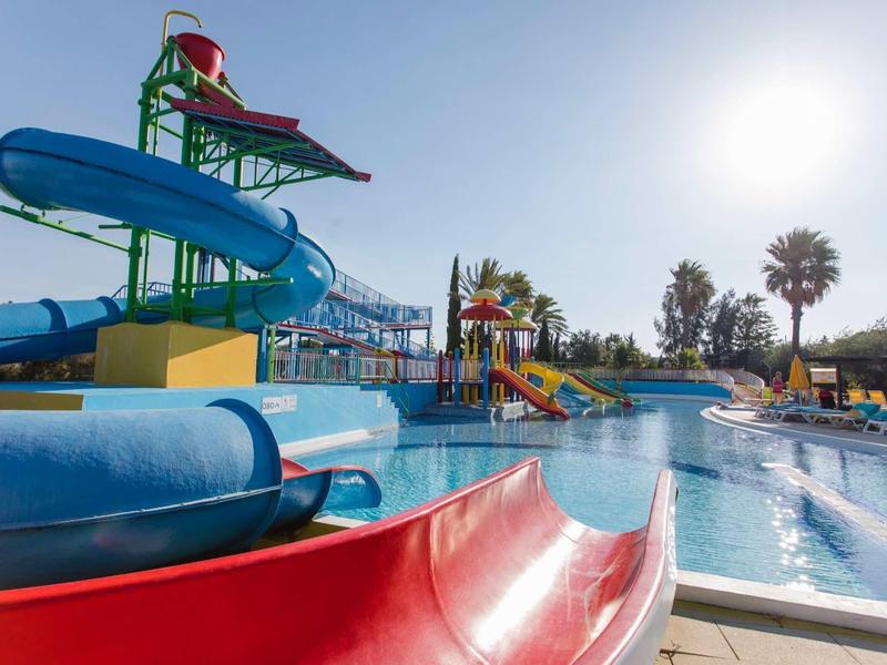 Glijbaan en zwembadgebied in een zonnig waterpark met palmbomen en blauwe lucht.