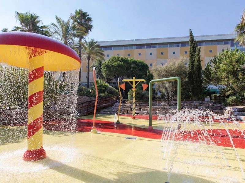 Kleurrijke waterspeeltuin met paddenstoelfontein en spuitend water onder blauwe lucht.