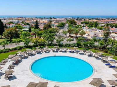 Groot rond zwembad omgeven door parasols en ligstoelen in een tuin met uitzicht op de stad.