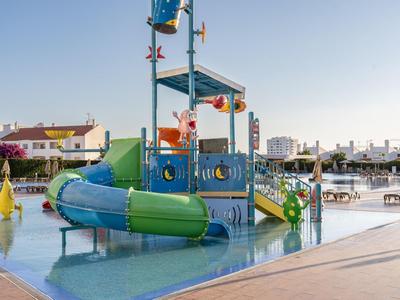 Kleurrijke waterspeeltuin met glijbanen in ondiep zwembad onder blauwe lucht en gebouwen.