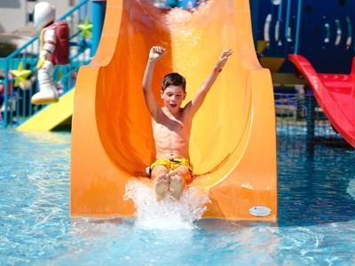 Kind glijdt vrolijk van een gele glijbaan het zwembad in.