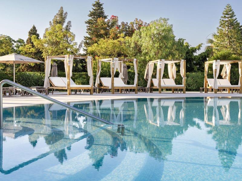 Heldere hotelzwembad met ligbedden en parasols in een groene tuin onder een blauwe lucht.