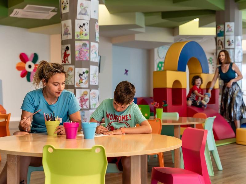Kinderen spelen en tekenen in een kleurrijke binnenruimte met speeltoestel en zitplaatsen.