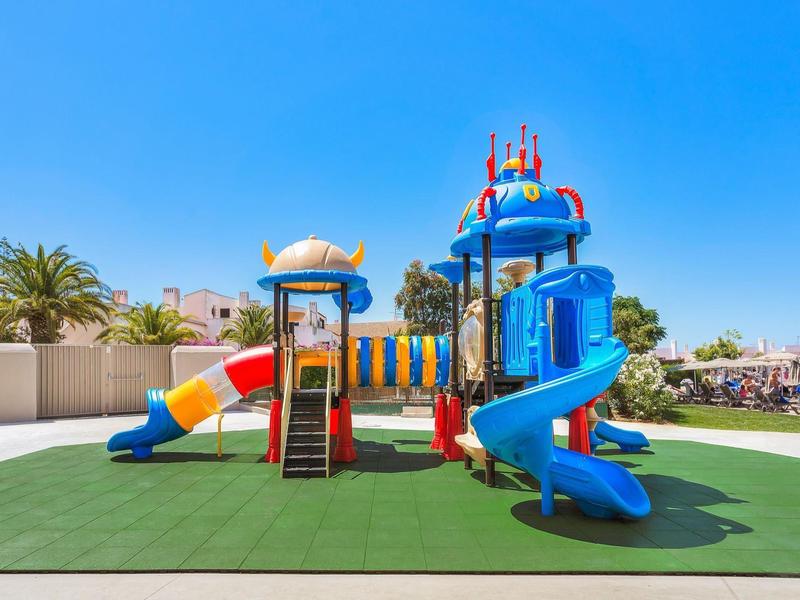 Kleurrijke speeltuin met glijbanen en klimnetten op groen gras onder een heldere hemel.