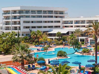 Groot resort met blauw zwembad, parasols en ligstoelen tussen palmbomen.