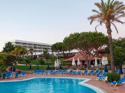 Buitenzwembad met ligstoelen en palmbomen voor een hotelgebouw bij zonsondergang.