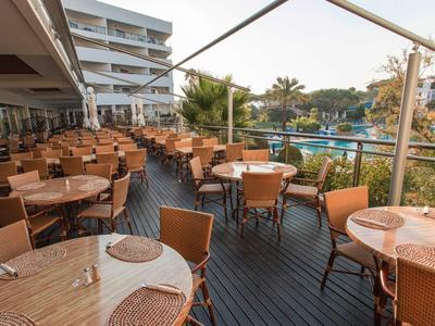 Terras met tafels en stoelen naast een hotel met uitzicht op een zwembad en palmbomen.