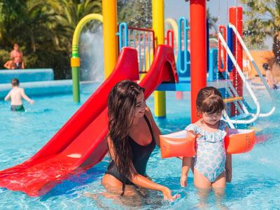 Vrouw en kind spelen in ondiep zwembad met rode glijbanen op de achtergrond.