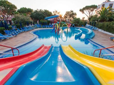 Kleurrijk zwembad met glijbanen, ligstoelen en bomen op de achtergrond.