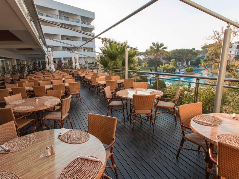 Terras met tafels en stoelen naast een hotel met uitzicht op een zwembad en palmbomen.