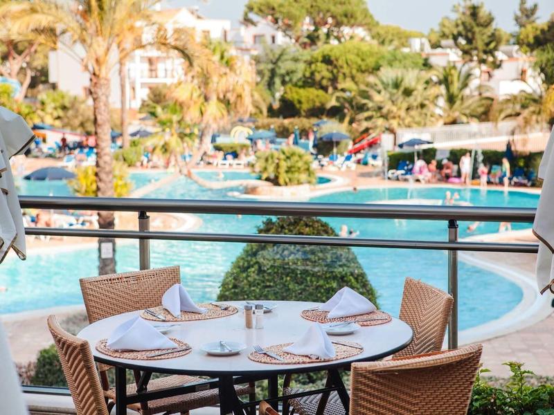 Een gedekte tafel met stoelen op een terras met uitzicht op een zwembad en palmbomen.