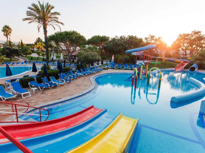 Buitenzwembad met kleurrijke glijbanen, ligstoelen en palmbomen bij zonsondergang.