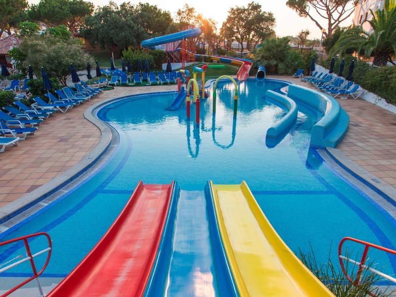 Kleurrijk zwembad met glijbanen bij een hotel met ligstoelen en groen.
