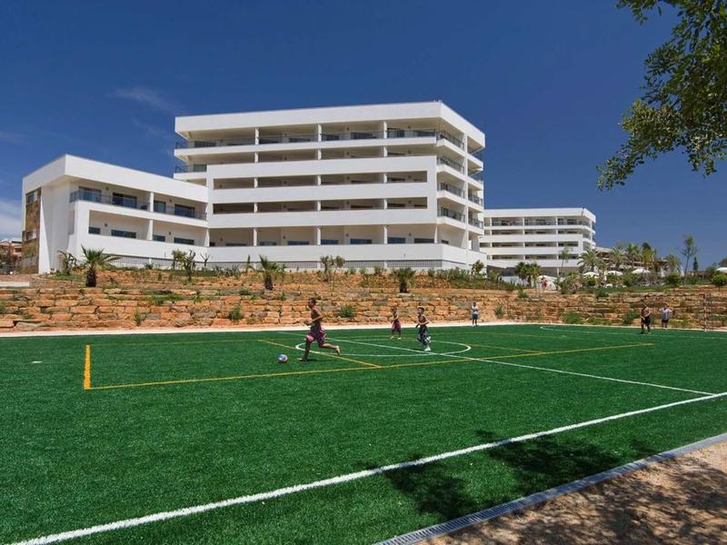 Modern hotelgebouw met meerdere verdiepingen achter een groen sportveld onder een heldere lucht.