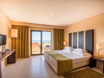 Helles Hotelzimmer mit Doppelbett, Fernseher und Balkon mit Meerblick.